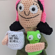 Close up photo. Crochet sculpture of Louise from Bob's Burgers, holding a mug of coffee that reads "I Will See You in Hell" and a green monster under her other arm.