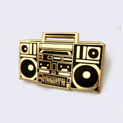 Gold enamel pin of a boombox, with black accents.