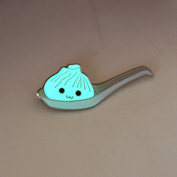 Enamel pin of an illustrated soup dumpling in a large ramen spoon. Dumpling has a smiling face. Pin is glowing in the dark.