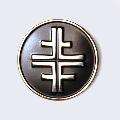Circular enamel pin, black with silver lining. In the center is a silver shape, the band Jawbreaker's logo. Logo is an elongated plus sign with four additional lines coming out the sides, with an intersection running through the middle and sides of the main shape.