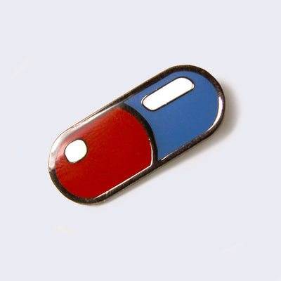 Enamel pin of a pill capsule, one side is red and the other is blue. Pin has a silver outline.