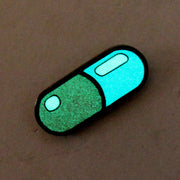 Enamel pin of a pill capsule, glowing in the dark.