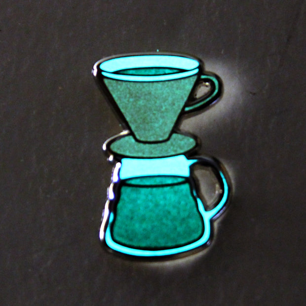 Enamel pin of a pour over coffee set, with an orange dripper and full pot of black coffee. Pin is glowing in the dark.