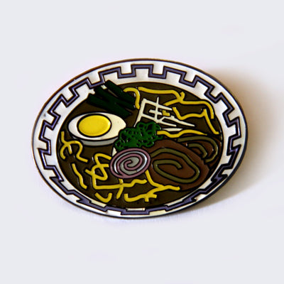 Enamel pin of a bowl of ramen. Ramen is brown and has an egg, fishcake, pork, seaweed and yellow noodles in the bowl.
