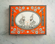 Fine line black ink illustration on bright orange panel, with the focal point encased in a decorative bordered oval in center of panel. 2 bunnies wearing dresses and flower crowns face each other and hold a connected chain of orange stars. Outside of the oval, are tassels on each side and decorative flowers on top and bottom.