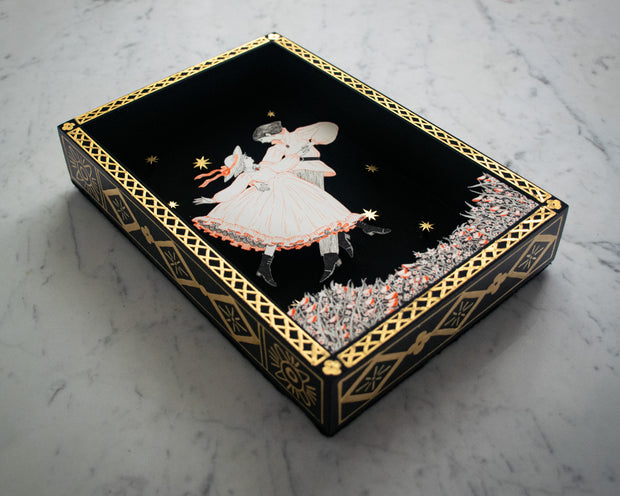 Shadowbox like illustration of a couple, dancing together over a bed of flowers. Illustration is done in very fine lines, orange and black on white paper. They are against an all black background with a gold pattern border and some gold stars floating. Side of the box has gold pattern detailing.