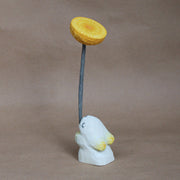 Whittled wooden sculpture of a small white bunny, with eyes closed and very simplistic body, facing away and holding a large whittled wooden yellow dandelion with a long green stem.