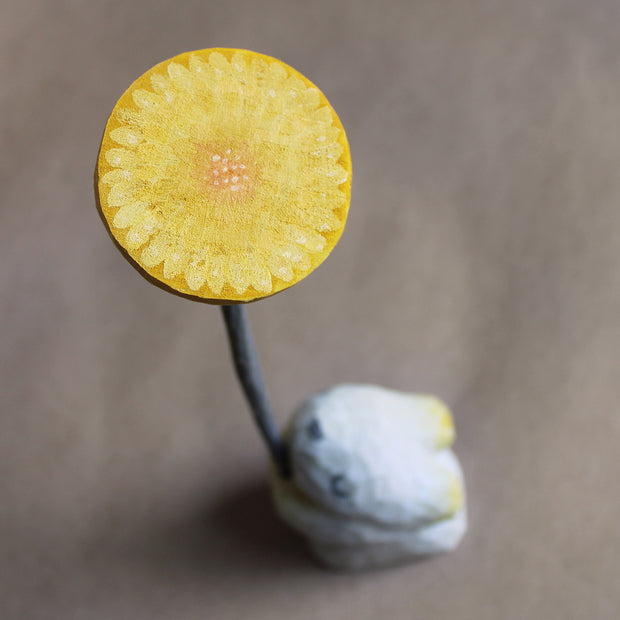 Aerial view of a whittled wooden dome shaped dandelion, with a dandelion painting on the flat surface. The dandelion has a long stem and is being held by a small white wooden bunny, which is out of focus.