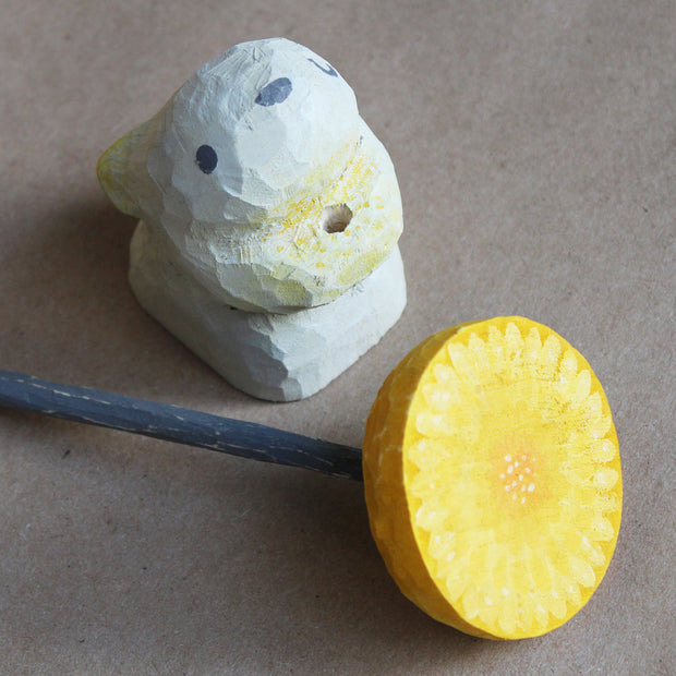 Whittled wooden sculpture of a small white bunny, with one eye closed and very simplistic body. It has a hole in its chest area, where a wooden dandelion can be placed. For now, the wooden whittled dandelion sits besides the bunny.