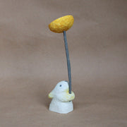 Whittled wooden sculpture of a small white bunny, with one eye opened and very simplistic body, facing away and holding a large whittled wooden yellow dandelion with a long green stem.