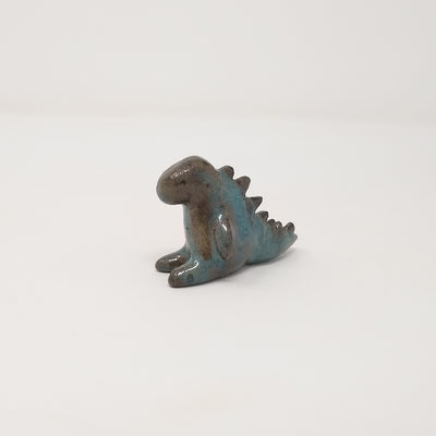 A small ceramic brown and blue Godzilla figure with minimal body and facial details, sitting on the ground with its legs extended in front and its tail extended behind. 