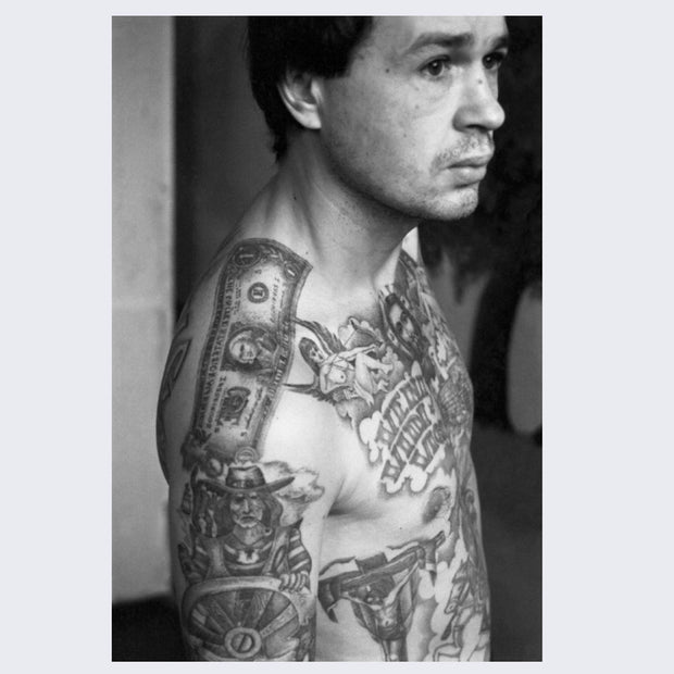 Page example, black and white photograph of a man looking off, various tattoos are displayed on his upper body.