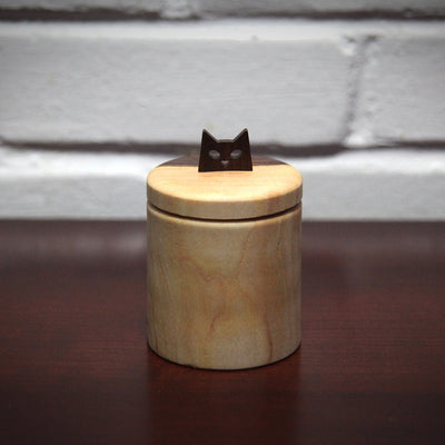 Cylindric wooden box, with a natural wood grain of light wood and darker exterior wood. As a handle for the top is a simplified cat head, with cut out eyes and a small nose.