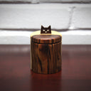 Cylindric wooden box, with a natural wood grain of light wood and darker exterior wood. As a handle for the top is a simplified cat head, with cut out eyes and a small nose.