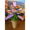 Sculpture of a plant made out of pink and blue risograph printed paper leaves, attached by wire to a pot. Leaves have a rounded striping pattern. It is in a wood grain pattern mug with many tufts of fake moss greenery.