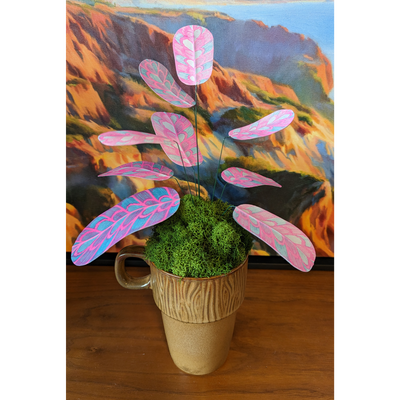 Sculpture of a plant made out of pink and blue risograph printed paper leaves, attached by wire to a pot. Leaves have a rounded striping pattern. It is in a wood grain pattern mug with many tufts of fake moss greenery.