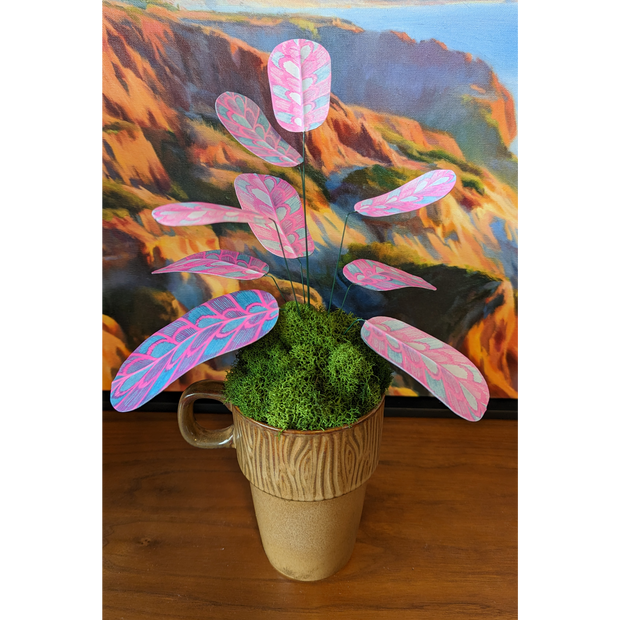 Sculpture of a plant made out of pink and blue risograph printed paper leaves, attached by wire to a pot. Leaves have a rounded striping pattern. It is in a wood grain pattern mug with many tufts of fake moss greenery.