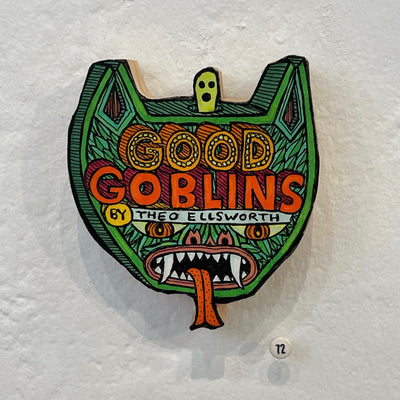 Illustrated woodcut green cat head. A small ghost pops out the top of the head, which has green fun, sharp teeth and a long red tongue. "Good Goblins by Theo Ellsworth" is written in a stylized font on the forehead.