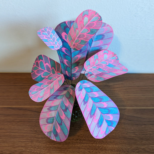 Sculpture of a plant made out of pink and blue risograph printed paper leaves, attached by wire to a pot. Leaves have a rounded striping pattern.