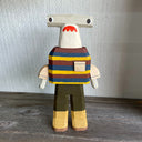 Sculpture made out of whittled wood and partially painted of a hammerhead shark wearing a striped shirt, pants and boots. The center of its body is very blocky.