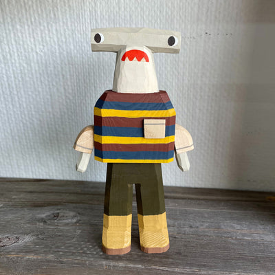 Sculpture made out of whittled wood and partially painted of a hammerhead shark wearing a striped shirt, pants and boots. The center of its body is very blocky.