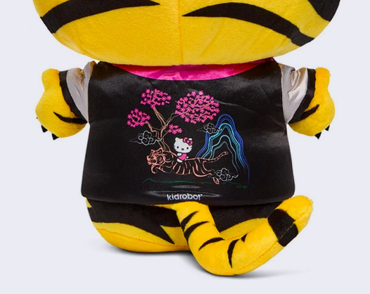 Back of Hello Kitty plush, wearing a full body tiger costume, that goes over her head like a hood. She also wears a black satin style jacket with a Japanese style illustration of a tiger, cherry blossom trees and Hello Kitty on the back of the jacket.