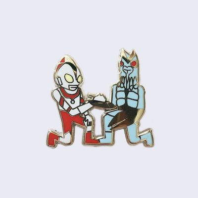 Enamel pin of two Japanese kaiju-esque figures kneeling and clasping hands with one another. One character is in a red and white armored suit and the other is a blue bug creature with claws.