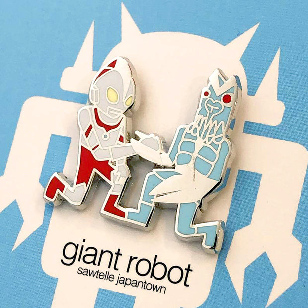 Close up of enamel pin of two Japanese kaiju-esque figures kneeling and clasping hands with one another.