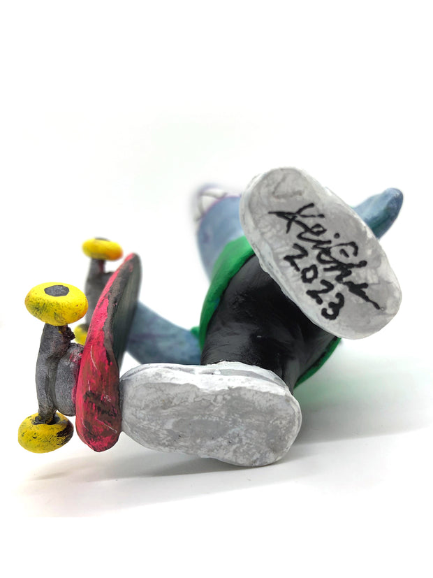 Sculpture of a cartoon style shark wearing a green t-shirt, jeans and white sneakers. He holds a red skateboard with a fishbone design and yellow wheels.