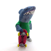 Sculpture of a cartoon style shark wearing a green t-shirt, jeans and white sneakers. He holds a red skateboard with a fishbone design and yellow wheels.