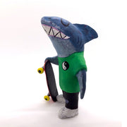 Sculpture of a cartoon style shark wearing a green t-shirt, jeans and white sneakers. He holds a red skateboard with a fishbone design and yellow wheels.