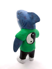 Sculpture of a cartoon style shark wearing a green t-shirt, jeans and white sneakers. He holds a red skateboard with a fishbone design and yellow wheels. He has a large yin yang pattern on the back of his shirt.