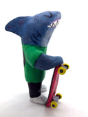 Sculpture of a cartoon style shark wearing a green t-shirt, jeans and white sneakers. He holds a red skateboard with a fishbone design and yellow wheels.