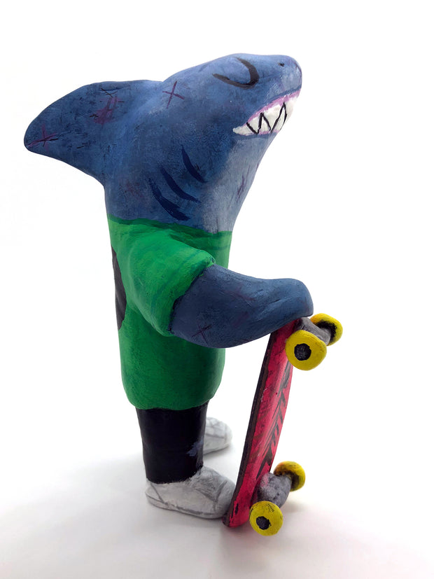 Sculpture of a cartoon style shark wearing a green t-shirt, jeans and white sneakers. He holds a red skateboard with a fishbone design and yellow wheels.