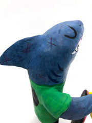 Sculpture of a cartoon style shark wearing a green t-shirt, jeans and white sneakers. He holds a red skateboard with a fishbone design and yellow wheels. His skin is very beat up with various scars.