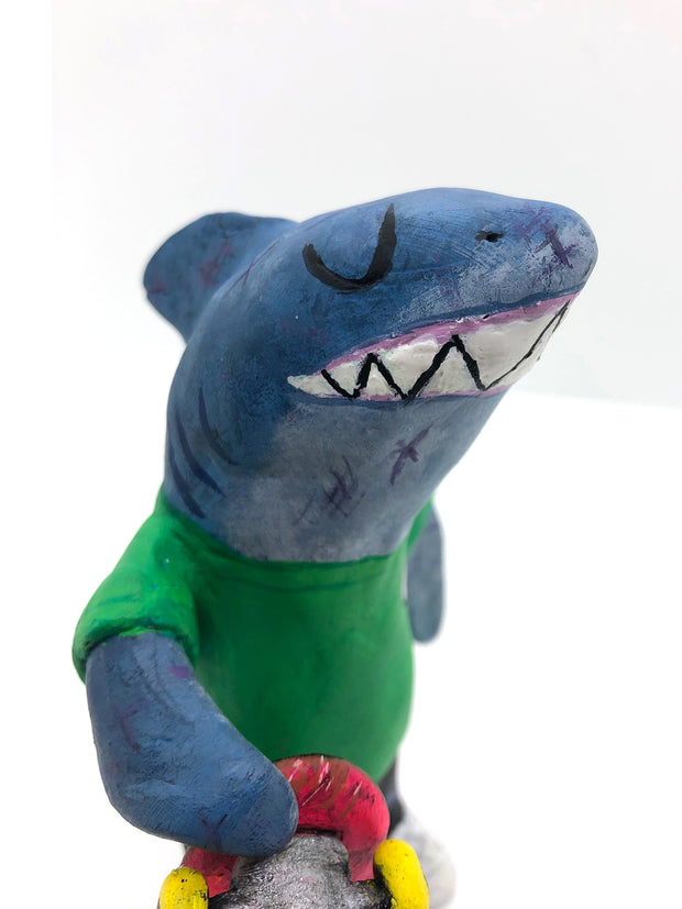 Sculpture of a cartoon style shark wearing a green t-shirt, jeans and white sneakers. He holds a red skateboard with a fishbone design and yellow wheels.