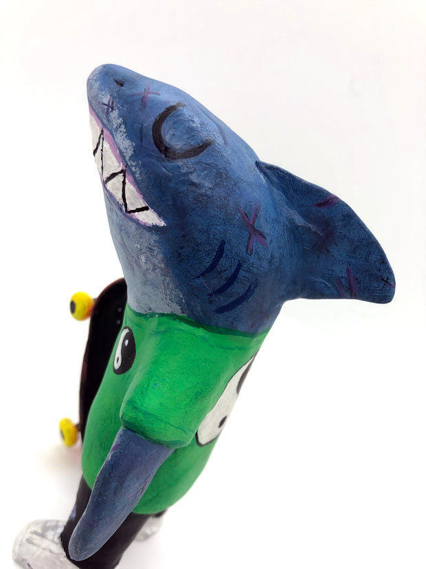 Sculpture of a cartoon style shark wearing a green t-shirt, jeans and white sneakers. He holds a red skateboard with a fishbone design and yellow wheels.