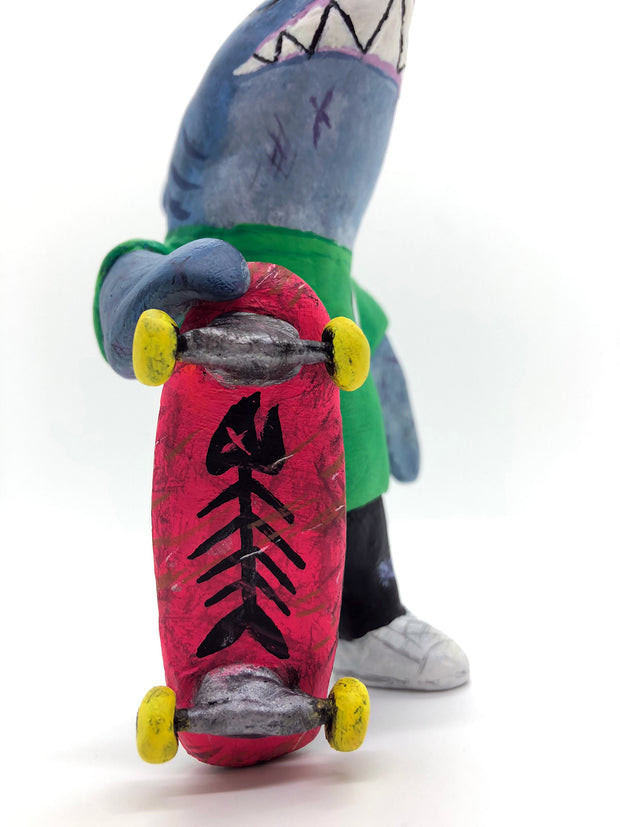 Sculpture of a cartoon style shark wearing a green t-shirt, jeans and white sneakers. He holds a red skateboard with a fishbone design and yellow wheels.