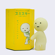 Simplified light green-yellow character, looking behind and holding both of its arms out in front of it. It stands next to its product box, with is all yellow with green Japanese script and an illustration of Smiski peeking around a corner.