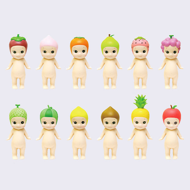 12 vinyl Kewpie babies, standing nude with different fruits atop their heads like hood hats. Options are: strawberry, peach, persimmon, pear, dragonfruit, grapes, melon, watermelon, lemon, durian, pineapple and apple.