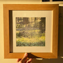 A hand holding a wooden frame with an intaglio print of a line art car that says "WOAHH" on the license plate and "Super Fast Car Club!!!" written below. The background is a photograph of a forest setting.