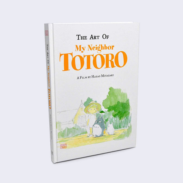 White hardcover book, with a small watercolor illustration of a young girl with a small Totoro and white chibi Totoro walking through a forest near a house. "The Art of My Neighbor Totoro" is written in logo font.