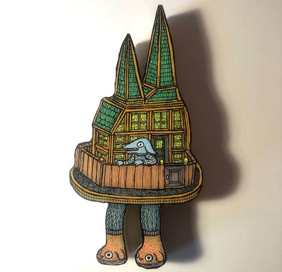 Die cut wooden sculpture of a tall Victorian style house, with a blue dog in a sweater and standing like a human, in the front yard. The yard has feet, standing under the house.