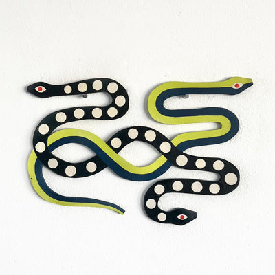 Die cut wood sculpture of two snakes intertwined with one another. One is green with teal stripe and the other is black with white polka dots and a head on each side.