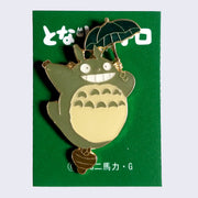Enamel pin of smiling Totoro balanced with one foot on an acorn and flying with a small, dark green umbrella.