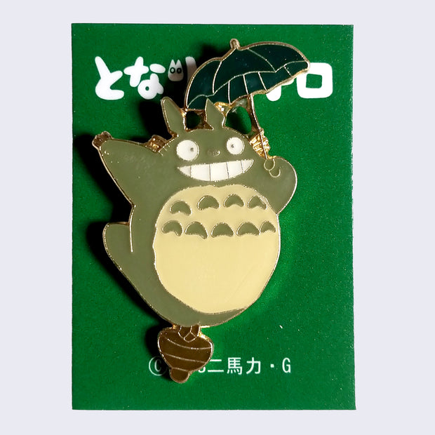 Enamel pin of smiling Totoro balanced with one foot on an acorn and flying with a small, dark green umbrella.