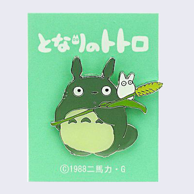 Enamel pin of a small Chibi-like Totoro standing with a surprised expression, looking to the side while holding a single willow. There is a small white Chibi Totoro on its shoulder.