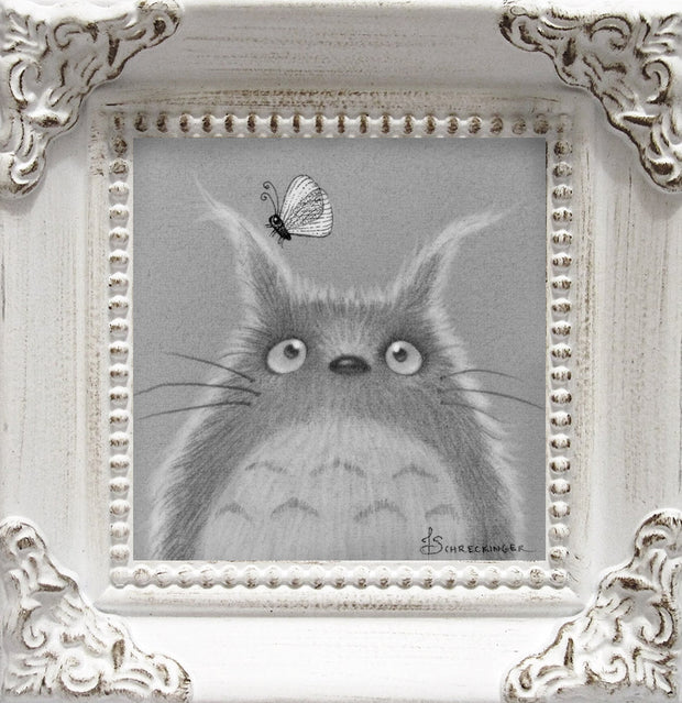 Soft textured graphite drawing of a slightly Chibi version of Totoro, looking up curiously at a butterfly on his ear, which is drawn in a more stark black line graphic style. Piece is framed in an ornate white wooden frame.