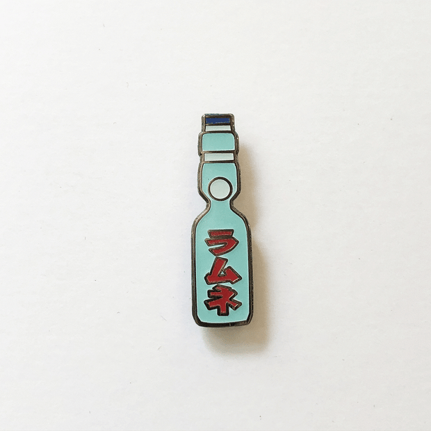 Gif of enamel pin of a Japanese Ramune Soft Drink, a light blue bottle with a white ball towards the top and red Japanese script down the middle. One image is of the pin in light, the other is of the pin glowing in the dark.