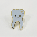 Gif of an enamel pin of an illustrated smiling white tooth with gold sparkles in the top right corner and a gold outline. One image is of the pin in light, the other is of the pin glowing in the dark.